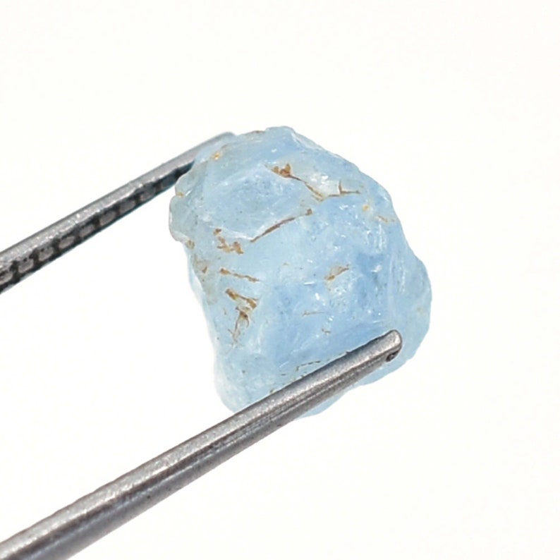 May include: A rough, light blue aquamarine gemstone held by tweezers. The gemstone has a natural, unpolished look.