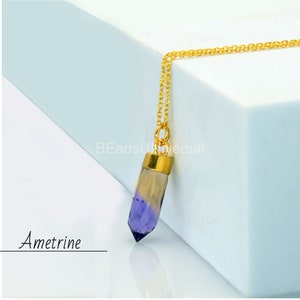 Peut inclure: Un collier en chaîne dorée avec un pendentif en cristal d'amétrine violet et jaune. Le pendentif est en forme de pointe et a un capuchon doré en haut. Le collier est sur une surface blanche. "Ametine" est écrit en noir sous le pendentif.