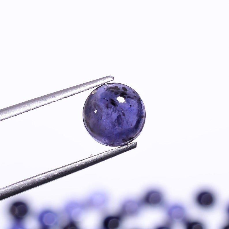 May include: A round, polished, deep blue gemstone is held by a pair of silver tweezers against a white background. The gemstone has dark inclusions and a smooth, reflective surface. Other similar gemstones are blurred in the background.