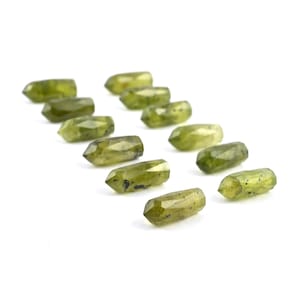May include: A collection of twelve small, faceted, olive-green gemstone points arranged in a staggered formation. Each individual stone features a double-terminated, elongated hexagonal shape with translucent surfaces and visible dark mineral inclusions.