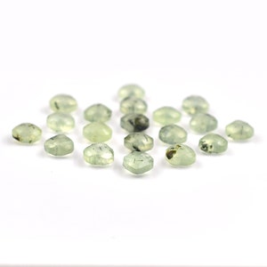 May include: A collection of small, faceted, pale green gemstones with visible dark inclusions. Each stone features a hexagonal shape with multiple polished surfaces that catch the light, displaying varying degrees of translucency and internal patterns.