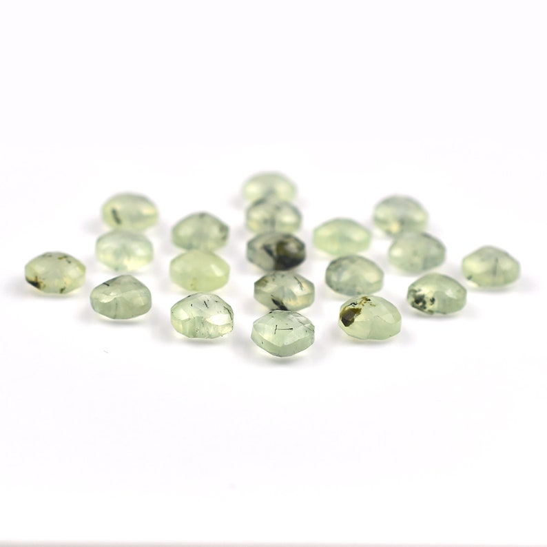 May include: A collection of small, faceted, pale green gemstones with visible dark inclusions. Each stone features a hexagonal shape with multiple polished surfaces that catch the light, displaying varying degrees of translucency and internal patterns.