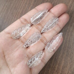 Può includere: Sei ciondoli a forma di foglia in cristallo di quarzo trasparente, ognuno intagliato con un design a foglia dettagliato. I ciondoli sono mostrati su una mano.