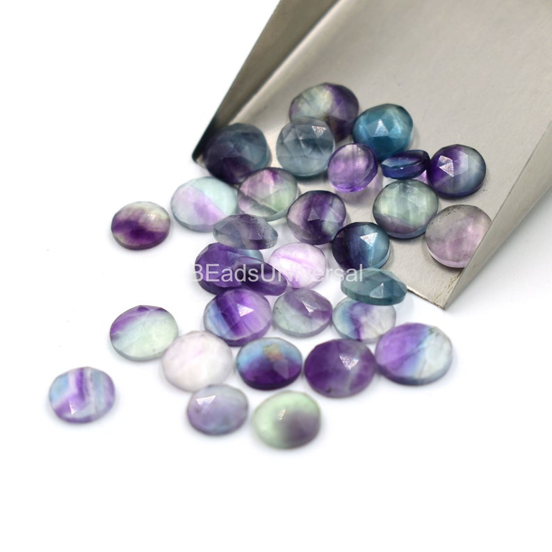 May include: A collection of round, faceted fluorite gemstones in shades of purple, green, and white. The stones are loose and scattered on a white surface.