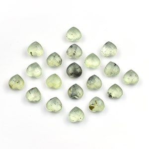 May include: A collection of seventeen small, heart-shaped gemstones in a pale, translucent green hue. Each stone features a faceted surface and visible dark, needle-like inclusions scattered throughout the light green mineral structure.