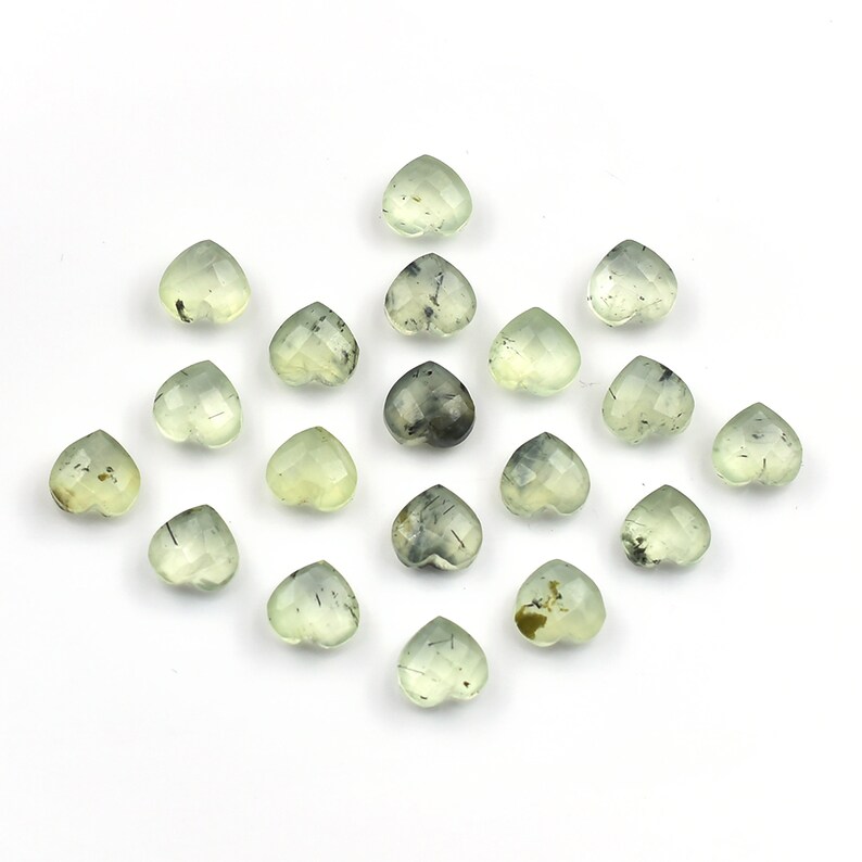 May include: A collection of seventeen small, heart-shaped gemstones in a pale, translucent green hue. Each stone features a faceted surface and visible dark, needle-like inclusions scattered throughout the light green mineral structure.