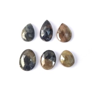 May include: A collection of six loose gemstones with faceted surfaces. The stones are arranged in two rows, featuring a mix of pear and oval shapes in various shades of dark blue, grey, and brownish-gold, displaying a polished finish.