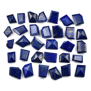 May include: A collection of faceted, deep blue lapis lazuli gemstones with gold flecks. The gemstones are cut into various geometric shapes, including rectangles, trapezoids, and diamonds. The stones are arranged on a white surface.
