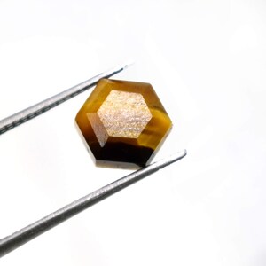 May include: A hexagonal tiger's eye gemstone, held by tweezers. The gemstone is a golden-brown colour with darker brown bands and a polished surface. The tweezers are positioned diagonally against a white background.