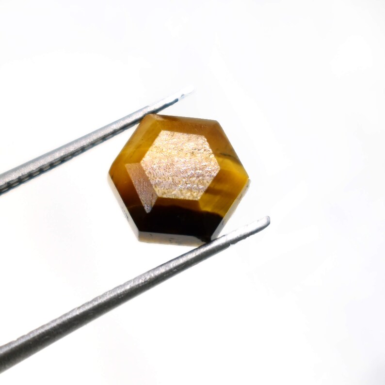 May include: A hexagonal tiger's eye gemstone, held by tweezers. The gemstone is a golden-brown colour with darker brown bands and a polished surface. The tweezers are positioned diagonally against a white background.