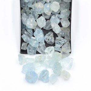 May include: A close-up of a pile of light blue aquamarine gemstones. The gemstones are rough and have a variety of shapes and sizes. The gemstones are spilling out of a black container.