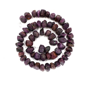 Könnte beinhalten: Eine Kette aus violetten Rubin-Edelsteinen, jede Perle hat eine raue, unregelmäßige Form. Die Perlen sind auf einem Silberdraht aufgefädelt.