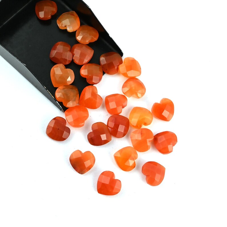 May include: A collection of small, faceted heart-shaped orange carnelian gemstones spilling out from a black metal scoop. The stones exhibit varying shades of translucent and opaque orange, ranging from light amber to deep reddish-orange tones.
