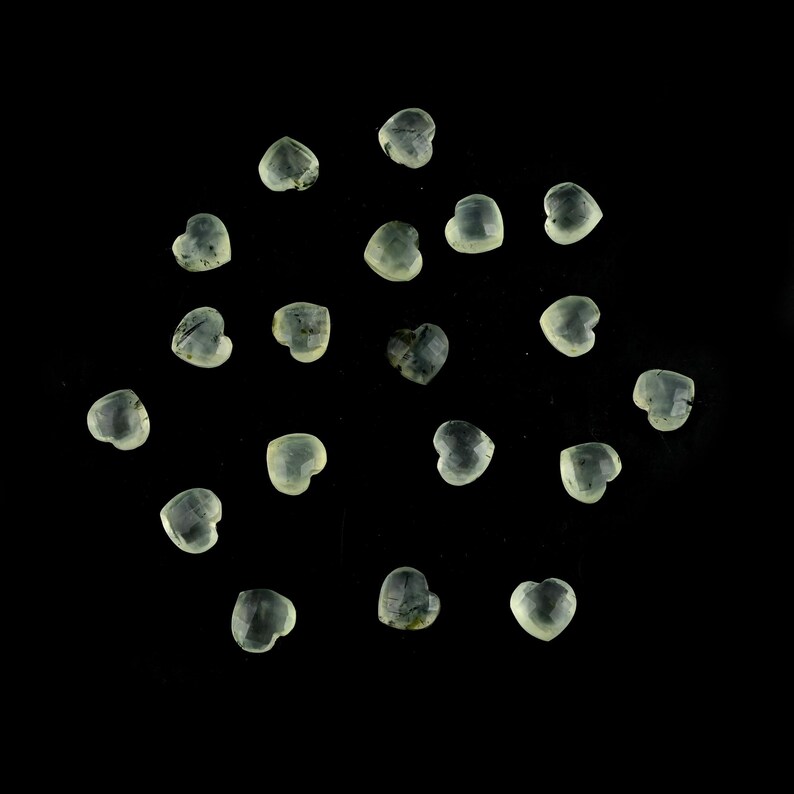 May include: A collection of twenty small, heart-shaped gemstones with a translucent, pale green hue. Each individual stone features a faceted surface that catches the light, displaying subtle internal inclusions and a smooth, polished finish.
