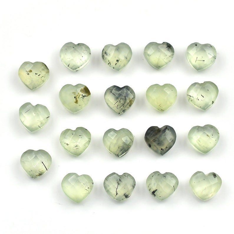 May include: A collection of nineteen heart-shaped gemstones with a faceted surface. Each stone is a pale, translucent light green color and features unique dark, needle-like inclusions scattered throughout the interior of the crystal structure.