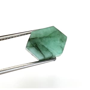 Könnte beinhalten: Ein grüner Smaragd mit sechseckiger Form und sichtbaren Einschlüssen. Der Smaragd wird von einer Pinzette gehalten.
