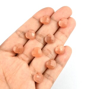 K&ouml;nnte beinhalten: Neun kleine, facettierte herzf&ouml;rmige Sonnenstein-Edelsteine mit einem durchscheinenden, pfirsich-orangen Farbton und einem subtilen inneren Schimmer. Diese polierten Steine weisen mehrere flache Oberfl&auml;chen auf, die das Licht einfangen.
