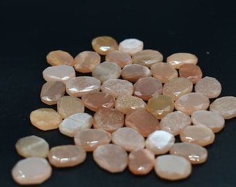 May include: A selection of faceted, oval gemstones in peach, tan, and cream hues. The stones are arranged on a dark backdrop, highlighting their polished surfaces and colour variations. These gemstones are suitable for jewellery making and crafts.
