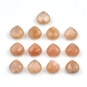 K&ouml;nnte beinhalten: Dreizehn facettierte herzf&ouml;rmige Perlen in einem sanften Pfirsich- oder Hellorangeton. Jede Perle weist eine geometrische, mehrfach facettierte Oberfl&auml;che auf, die das Licht reflektiert und in einem Raster auf wei&szlig;em Hintergrund angeordnet ist.