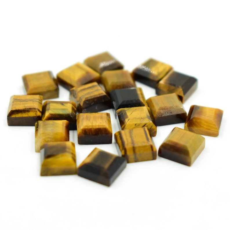 May include: A selection of square-cut tiger's eye gemstones. The stones exhibit a blend of golden brown and dark brown tones, featuring natural striped patterns. The gemstones are presented on a white surface, highlighting their polished facets.