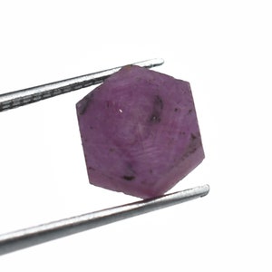 Könnte beinhalten: Ein rosafarbener, facettierter, sechseckiger Rubin-Edelstein mit Einschlüssen, gehalten von einer Pinzette.