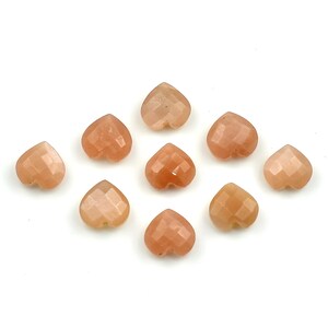 K&ouml;nnte beinhalten: Neun facettierte herzf&ouml;rmige Perlen in einem sanften Pfirsichton. Jede Perle weist eine geometrisch geschliffene Oberfl&auml;che auf, die das Licht reflektiert und ein dezentes Schimmern auf dem Stein erzeugt. Die Perlen sind locker auf wei&szlig;em Grund angeordnet.