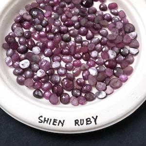May include: A collection of faceted, deep red-purple Shien Ruby gemstones displayed on a white plate. The gemstones vary in size and shape, with a mix of light and dark tones. The plate has the words "SHIEN RUBY" printed on it.