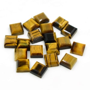 May include: A collection of square-cut tiger's eye gemstones. The stones are a mix of golden brown and dark brown, with natural patterns. The gemstones are scattered on a white surface, showcasing their polished facets.