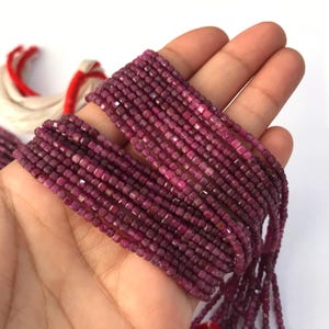 May include: A collection of multiple strands of small, faceted, deep magenta-colored square beads. The beads are strung together in long, thin lines, showcasing a uniform, geometric shape and a rich, saturated pinkish-purple hue throughout.