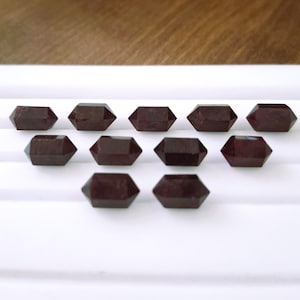 May include: Ten dark red, faceted, double-pointed gemstone crystals. The crystals are arranged in two rows on a white surface.