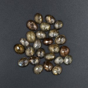 May include: A collection of 18 faceted oval-shaped gemstones in shades of brown and gray. The stones have a rough, natural finish and are arranged in a loose pile.