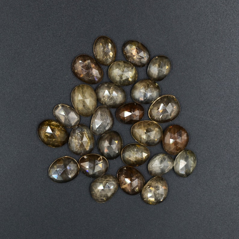 May include: A collection of 18 faceted oval-shaped gemstones in shades of brown and gray. The stones have a rough, natural finish and are arranged in a loose pile.