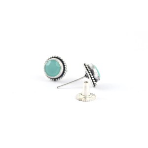 May include: A pair of round stud earrings with a light blue gemstone center and a silver-colored, rope-like border. One earring is detached, showing the post and backing. The earrings are set against a white background.