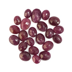 May include: A collection of twenty-one oval-shaped, faceted ruby gemstones in varying shades of deep pink and red. Each stone features a polished surface with geometric facets that reflect light, arranged in a loose cluster against a white background.