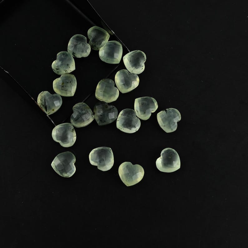 May include: A collection of small, faceted heart-shaped gemstones in a pale, translucent green hue. Each stone features a polished surface with subtle internal dark inclusions, scattered across the frame with some spilling from a black container.