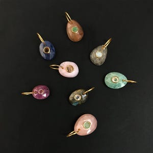 Puede incluir: Una colección de ocho colgantes de forma ovalada, cada uno con una piedra preciosa facetada engastada en un bisel de metal dorado con una anilla a juego. Los colgantes presentan varios colores, incluyendo tonos azules, rosas, verdes y grises.