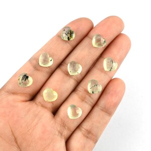 May include: Eight small, heart-shaped gemstones with a pale yellow-green hue and visible dark, needle-like inclusions. Each stone features a faceted surface that reflects light, arranged across the palm of a hand for scale and display.