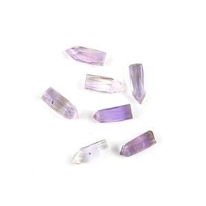 May include: A collection of seven small, faceted amethyst crystal points. Each piece features a translucent, light purple hue with varying degrees of clarity and natural internal inclusions, shaped into elongated, pointed geometric forms.