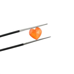 May include: A small, faceted, heart-shaped orange gemstone held by a pair of thin, silver-toned metal tweezers. The stone features a vibrant, translucent orange hue with multiple geometric facets that reflect light across its surface.