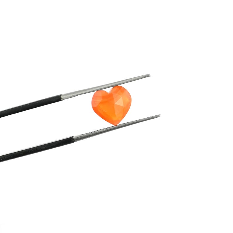 May include: A small, faceted, heart-shaped orange gemstone held by a pair of thin, silver-toned metal tweezers. The stone features a vibrant, translucent orange hue with multiple geometric facets that reflect light across its surface.