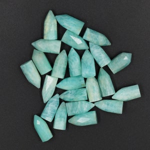 Puede incluir: Una colección de veintiuna piedras preciosas de amazonita pequeñas, pulidas y de color azul verdoso claro, cada una con forma de bala o punta facetada. Las piedras muestran variaciones de color naturales e inclusiones blancas en un grupo disperso.