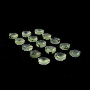 May include: A collection of fifteen faceted, pale green gemstones arranged in a grid. Each translucent stone features distinct, thin black needle-like inclusions scattered throughout the light mint-colored surface, creating a unique pattern.