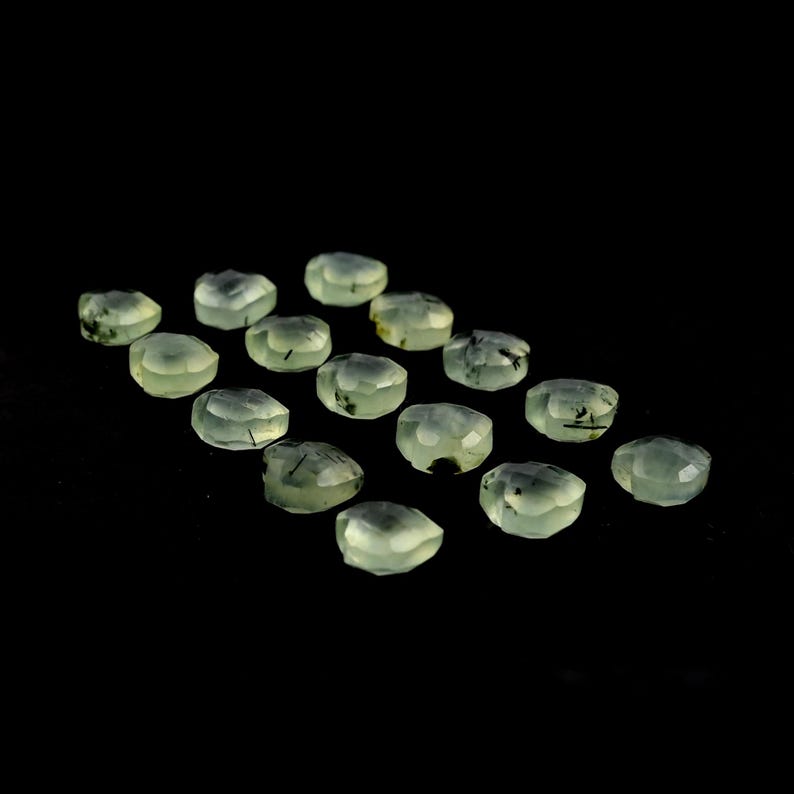 May include: A collection of fifteen faceted, pale green gemstones arranged in a grid. Each translucent stone features distinct, thin black needle-like inclusions scattered throughout the light mint-colored surface, creating a unique pattern.