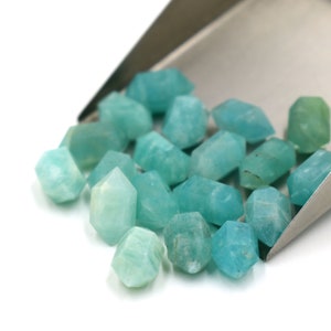 May include: A group of teal green, faceted, gemstone beads in a metal scoop. The beads are all roughly the same size and shape, and they have a smooth, polished finish.