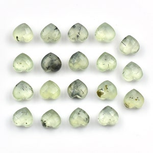 May include: A collection of seventeen heart-shaped gemstones arranged in a grid. Each stone features a faceted surface and a pale, translucent green color with visible dark, needle-like inclusions scattered throughout the crystalline structure.