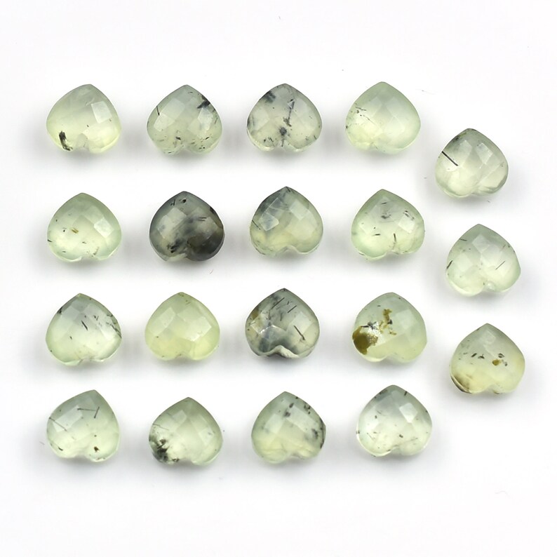 May include: A collection of seventeen heart-shaped gemstones arranged in a grid. Each stone features a faceted surface and a pale, translucent green color with visible dark, needle-like inclusions scattered throughout the crystalline structure.