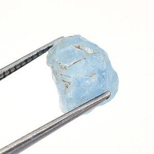 May include: A rough, light blue aquamarine gemstone held by tweezers. The gemstone has a natural, unpolished look.