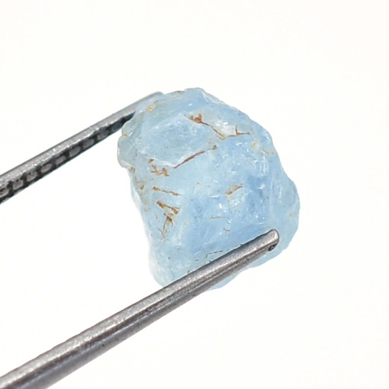 May include: A rough, light blue aquamarine gemstone held by tweezers. The gemstone has a natural, unpolished look.