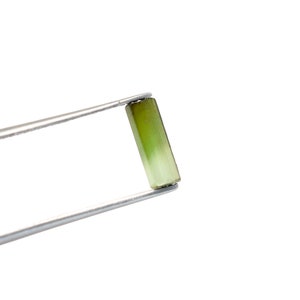 May include: A rectangular, light green gemstone held by silver tweezers. The gemstone has a gradient of color, transitioning from a darker green at one end to a lighter shade. The background is plain white.