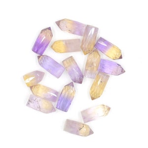 May include: A collection of fifteen small, faceted crystal points arranged in a scattered pattern. Each piece features a distinct color gradient, transitioning from a translucent purple amethyst tone at the base to a pale yellow citrine tip.